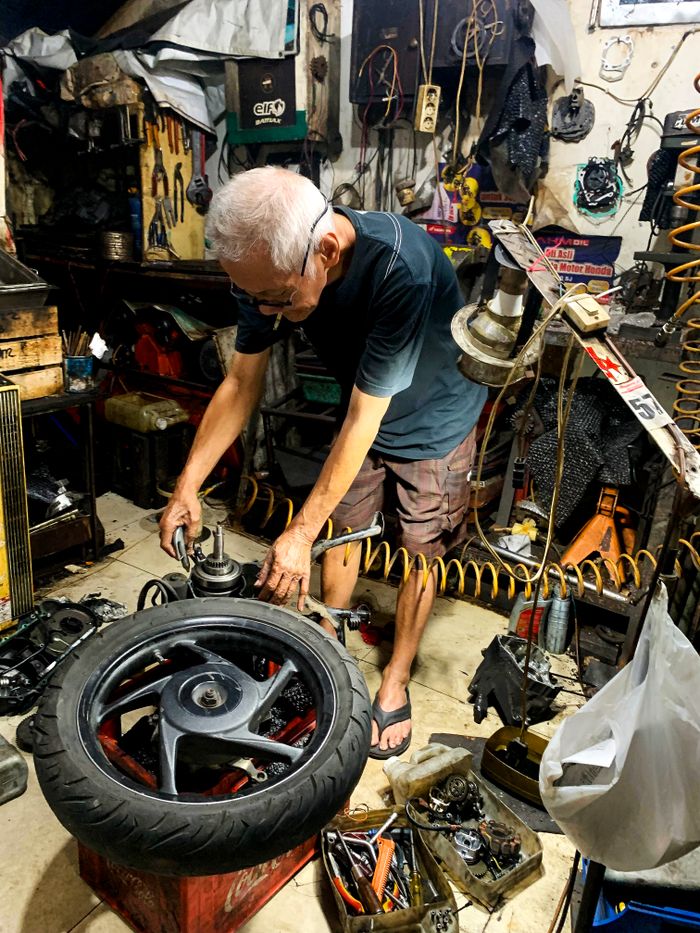 Elderly mechanic repairing a motorcycle tire in a cluttered workshop in Bandung, Indonesia.